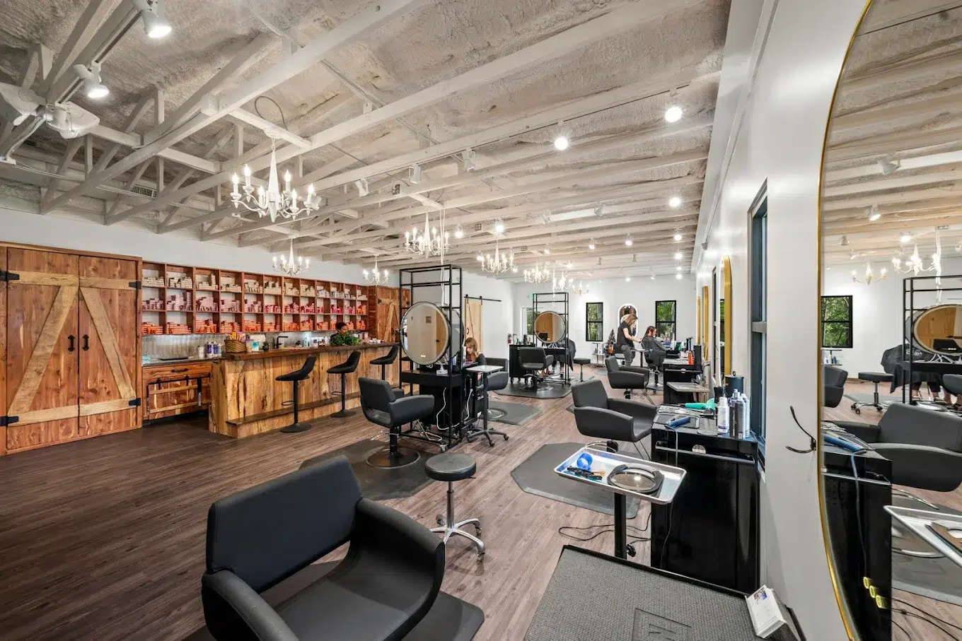 Modern rustic hair salon interior with styling stations, round mirrors, wooden product wall and chandeliers